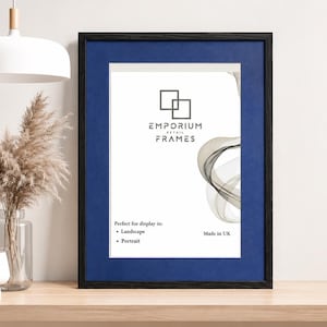 May include: A black picture frame with a blue mount, featuring the Emporium Retail Frames logo. The frame is on a wooden shelf, with a white lamp and vase. Designed for landscape and portrait display, Made in UK.