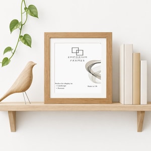May include: A light oak picture frame with the text "EMPORIUM FRAMES" and a graphic. The frame is on a wooden shelf with a wooden bird sculpture and books. A green plant hangs from above. Made in UK.