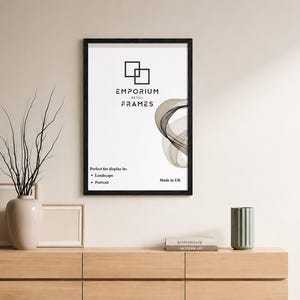 May include: A black framed print from Emporium Retail Frames hangs on a cream wall. The print features the company logo and abstract design. A wooden cabinet below holds a vase with branches, two smaller frames, and a metal container.