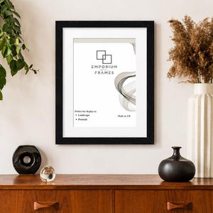 May include: A black picture frame with a white mount, featuring the Emporium Frames logo. The frame sits on a wooden cabinet, alongside a white vase with dried plants. A black ornament and a glass sphere are also on the cabinet.