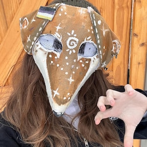 May include: A handmade fox mask with a brown and white color scheme. The mask features white painted details, mesh eye coverings, and a non-binary pride flag. The mask is worn over a green knit hat.