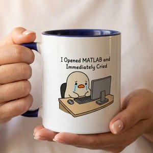 May include: A white ceramic mug with a dark blue interior and handle. The mug features a cartoon illustration of a crying character sitting at a desk with a computer and the text "I Opened MATLAB and Immediately Cried" printed in black.