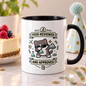 May include: A white ceramic mug with a black interior and handle. The front features a graphic of a grey and white cat wearing sunglasses and dabbing while holding laboratory flasks. Text reads: Seal of Review, PEER REVIEWED, AND APPROVED.
