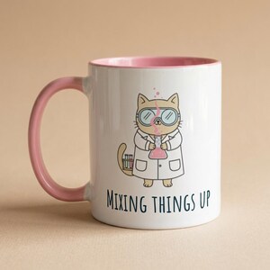 Peut inclure: Mug en céramique blanche avec intérieur et anse roses. Le mug présente un chat de dessin animé portant des lunettes de laboratoire et une blouse blanche, tenant une fiole. Le texte "MIXING THINGS UP" est imprimé en dessous.