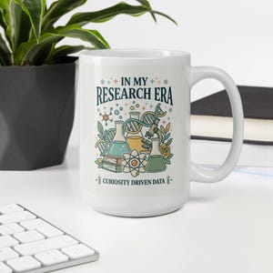 May include: White ceramic mug with a science-themed design. The mug features the text "IN MY RESEARCH ERA" above illustrations of beakers, a DNA strand, and an atom. The phrase "CURIOSITY DRIVEN DATA" is printed below the illustrations.