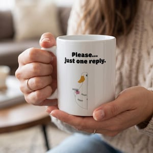 以下が含まれることがあります： 「Please... just one reply」の文字が入った白いセラミックマグ。 銃を持った漫画のアヒルのイラストが側面にプリントされています。 マグカップは人が持っています。