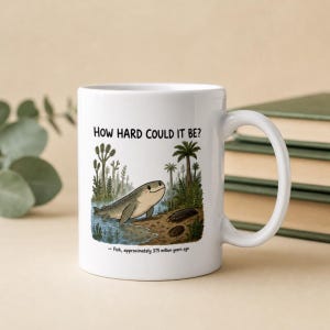 Puede incluir: Una taza de cerámica blanca con una ilustración de dibujos animados de un pez prehistórico saliendo del agua a la tierra. El texto "HOW HARD COULD IT BE?" aparece arriba y "- Fish, approximately 375 million years ago" abajo, con plantas verdes.