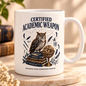 Peut inclure: Un mug en céramique blanc orné d'un hibou perché sur une pile de livres intitulés Biology, Physics, Data et Philosophy. Le texte indique Certified Academic Weapon et Becoming What Knowledge Demands, avec des illustrations d'une sphère armillaire.