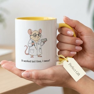 Puede incluir: Taza de cerámica blanca con interior y asa amarillos. La taza presenta un ratón de dibujos animados con gafas y bata de laboratorio, sosteniendo una jeringa y un paquete de pastillas. El texto en la taza dice: "¡Funcionó la última vez, lo juro!" Una etiqueta de regalo dice "Regalo perfecto".
