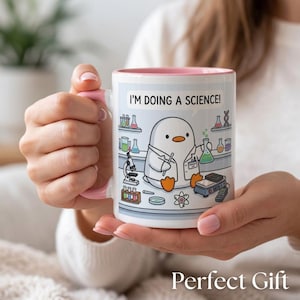 Peut inclure: Un mug en céramique blanc avec une anse et un intérieur roses, illustré d'un pingouin en blouse de laboratoire tenant un bécher. Le texte I'M DOING A SCIENCE! est inscrit au-dessus, entouré d'équipements de laboratoire comme des tubes à essai.