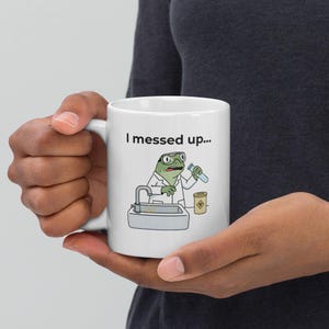 May include: White ceramic mug featuring a cartoon frog in a lab coat, holding a test tube. The frog is standing in front of a sink with a hazardous waste symbol. The text "I messed up..." is printed above the frog.