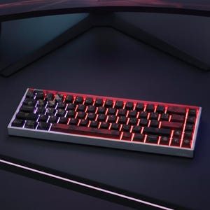 Może przedstawiać: Kompaktowa, podświetlana klawiatura mechaniczna ze srebrną ramką. Czarne klawisze mają marmurkowy wzór, a klawisze są podświetlane czerwonym i fioletowym światłem. Klawiatura znajduje się na ciemnej powierzchni, w tle monitor komputera.