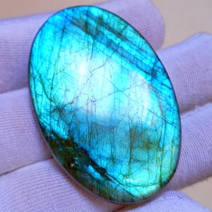 Puede incluir: Gema ovalada de labradorita con una vibrante exhibición de iridiscencia azul, turquesa y verde. La piedra pulida tiene una superficie lisa con inclusiones y grietas naturales, sobre un fondo blanco.