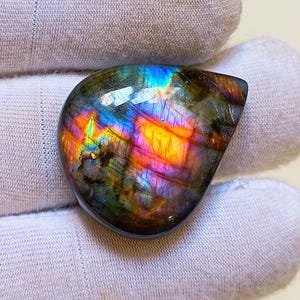Puede incluir: Una piedra preciosa de labradorita pulida e iridiscente. La piedra muestra un vibrante juego de colores, con destellos de azul, morado, naranja y amarillo. La gema tiene una forma suave y redondeada, sobre un fondo blanco.