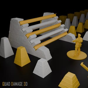 Könnte beinhalten: Ein 3D-gedrucktes Miniatur-Militärgeländeset mit einer Betonbarriere mit gelben Balken, pyramidenförmigen Hindernissen in Gelb und Grau und einer kleinen Soldatenfigur. Der Text "QUAD DAMAGE 3D" befindet sich unten.