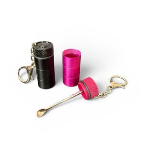 Puede incluir: Un juego de recipientes metálicos negros y rosas con una cuchara pequeña. El juego incluye un recipiente negro con una parte superior texturizada, un recipiente rosa y un recipiente rosa con una cuchara adjunta. Todas las piezas tienen llaveros.