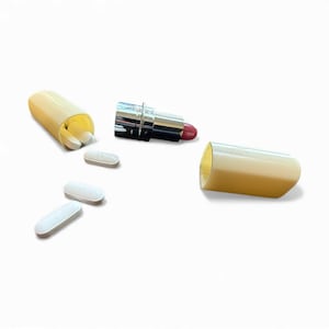 Peut inclure: Un contenant en forme de rouge à lèvres avec un rouge à lèvres rouge et plusieurs pilules blanches éparpillées. Le contenant est ouvert, révélant les pilules à l'intérieur. Le rouge à lèvres a une finition métallique argentée.