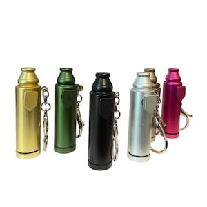 May include: A collection of colorful, cylindrical keychain accessories in gold, green, black, silver, and pink. Each has a small, rectangular clasp and a keyring. The items are arranged against a white background.