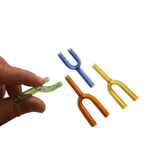 May include: A collection of colorful glass Y-shaped pipes in green, blue, orange, and yellow. One pipe is held by a hand, while the others are arranged on a white background. The pipes have a rounded shape.