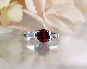 May include: A silver ring with a red gemstone and two smaller clear gemstones on either side.
