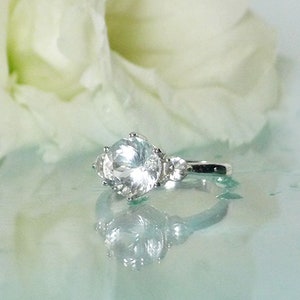 May include: A silver ring with a large, clear, round gemstone surrounded by smaller, clear gemstones. The ring is on a light blue surface with a white flower in the background.