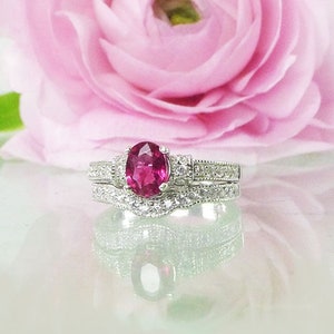May include: A silver ring set with a pink oval gemstone surrounded by smaller clear gemstones. The ring is on a white surface with a pink flower behind it.