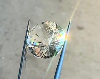 Puede incluir: Una gema transparente y facetada sostenida por unas pinzas. La gema es redonda y tiene un patrón de estrella en el centro.