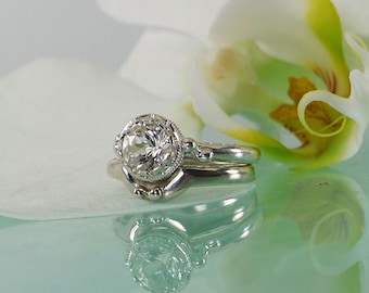May include: A set of two silver wedding bands with a large, round, clear gemstone in the center of the top band. The bands are on a white flower petal.