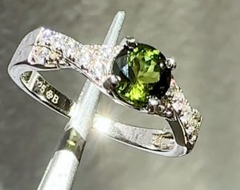 May include: A silver ring featuring a round, vibrant green gemstone as the focal point. The band is adorned with small, sparkling clear stones. The ring is held by a tool, showcasing the intricate design and craftsmanship.