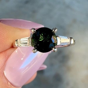 May include: A silver ring with a round, dark green gemstone surrounded by smaller, clear gemstones.