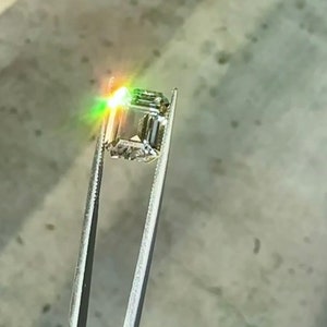 May include: A clear, square-shaped gemstone with a slight green hue is held by tweezers. The gemstone is reflecting light, creating a rainbow effect.
