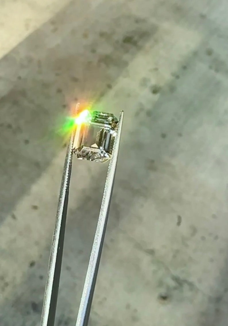 May include: A clear, square-shaped gemstone with a slight green hue is held by tweezers. The gemstone is reflecting light, creating a rainbow effect.