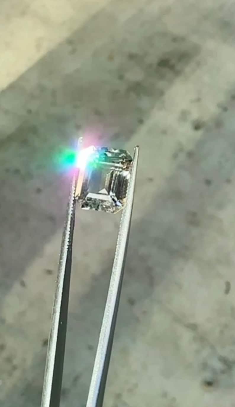 May include: A clear, square-shaped gemstone held by tweezers. The gemstone is reflecting rainbow colors.