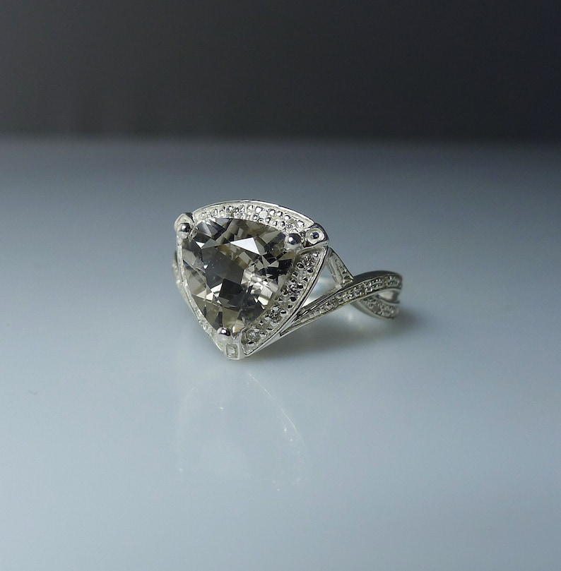 K&ouml;nnte beinhalten: Ein silberner Ring mit einem gro&szlig;en, dreieckigen, grauen Edelstein, der von kleineren, klaren Edelsteinen umgeben ist. Das Band ist verdreht und hat ein filigranes Design.