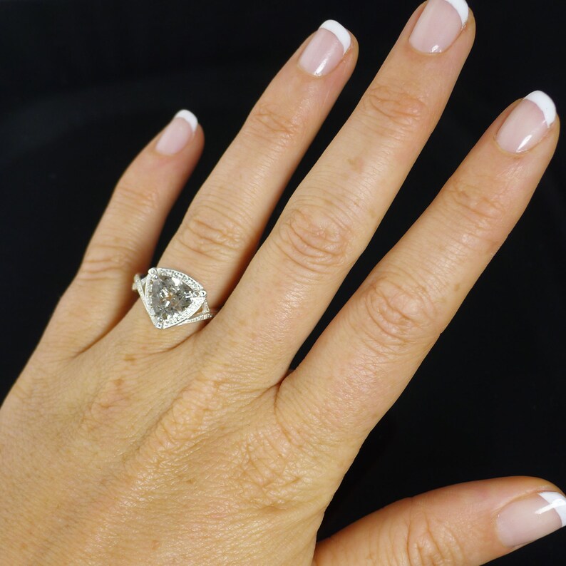 K&ouml;nnte beinhalten: Ein silberner Ring mit einem gro&szlig;en, klaren, dreieckigen Edelstein, der von kleineren Diamanten umgeben ist. Der Ring wird an einem Finger getragen.