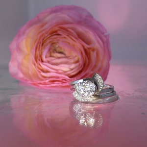 Könnte beinhalten: Ein silberner Verlobungsring mit einem großen, runden, klaren Diamanten, der von kleineren Diamanten umgeben ist. Der Ring liegt auf einem rosa Hintergrund mit einer rosa Blume.