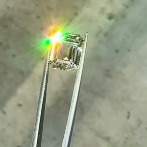 May include: A clear, square-shaped gemstone with a slight green hue is held by tweezers. The gemstone is reflecting light, creating a rainbow effect.