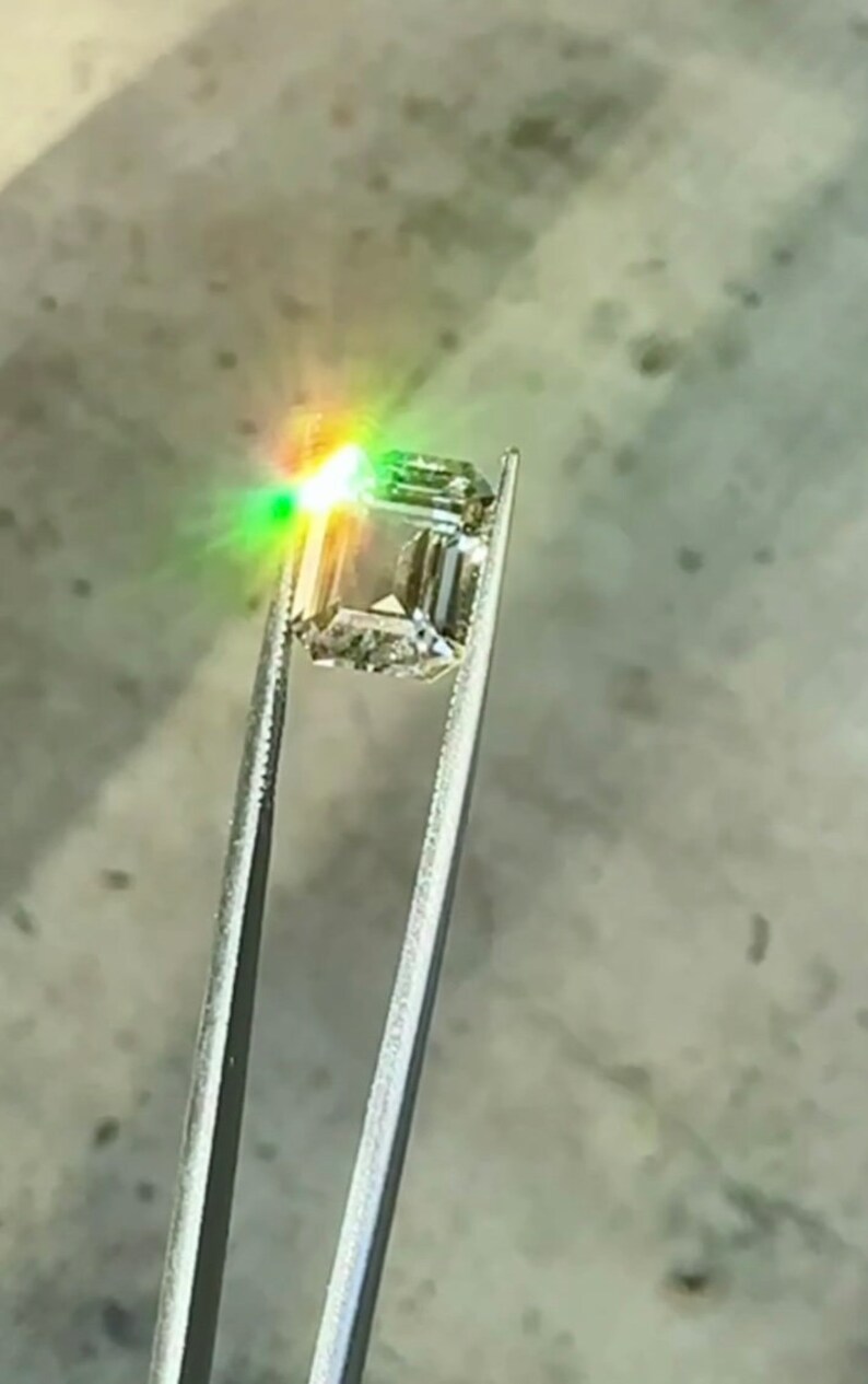 May include: A clear, square-shaped gemstone with a slight green hue is held by tweezers. The gemstone is reflecting light, creating a rainbow effect.