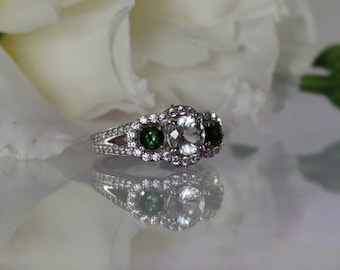 May include: A silver ring with a large, clear center stone surrounded by smaller diamonds. Two smaller green stones are set on either side of the center stone.