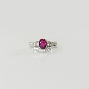 May include: A set of two silver rings with a large oval ruby gemstone in the center of the top ring. The bottom ring is a delicate band with small diamonds.