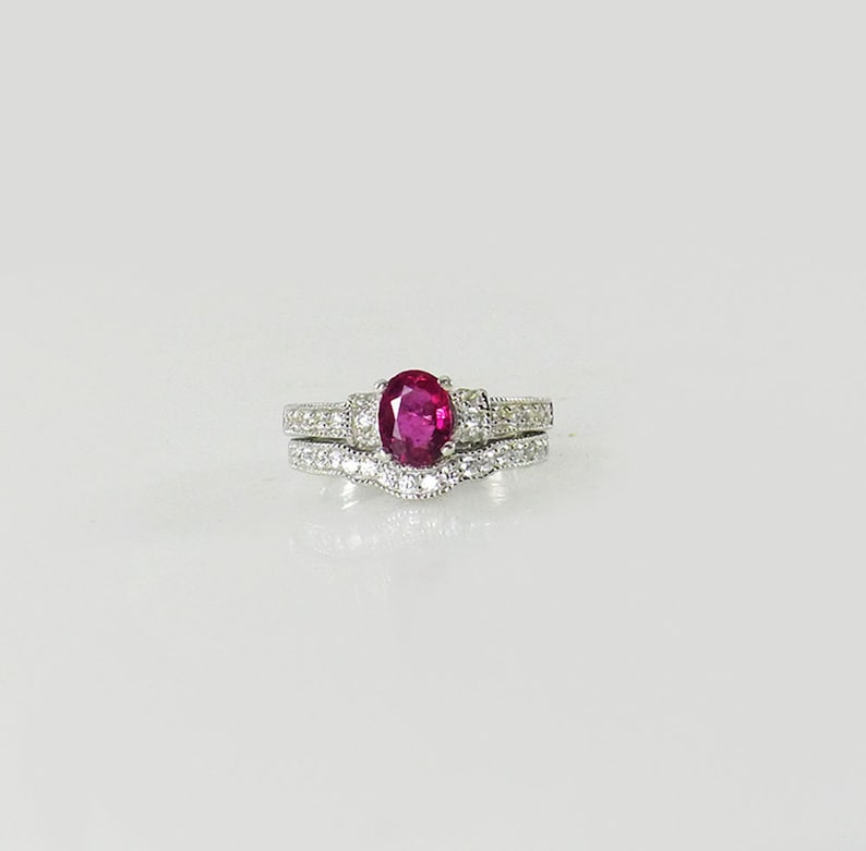 May include: A set of two silver rings with a large oval ruby gemstone in the center of the top ring. The bottom ring is a delicate band with small diamonds.