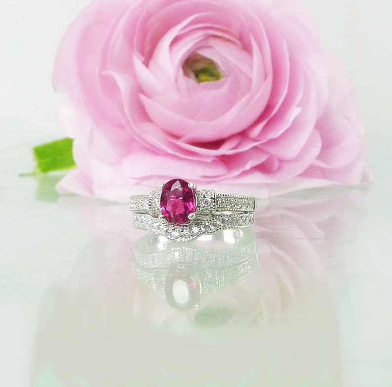 May include: A silver ring set with a pink oval gemstone surrounded by smaller clear gemstones. The ring is on a white surface with a pink flower behind it.
