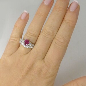 May include: A close-up of a woman's hand wearing a ruby and diamond engagement ring set. The ring features a large oval-shaped ruby surrounded by a halo of smaller diamonds. The band is also set with diamonds.