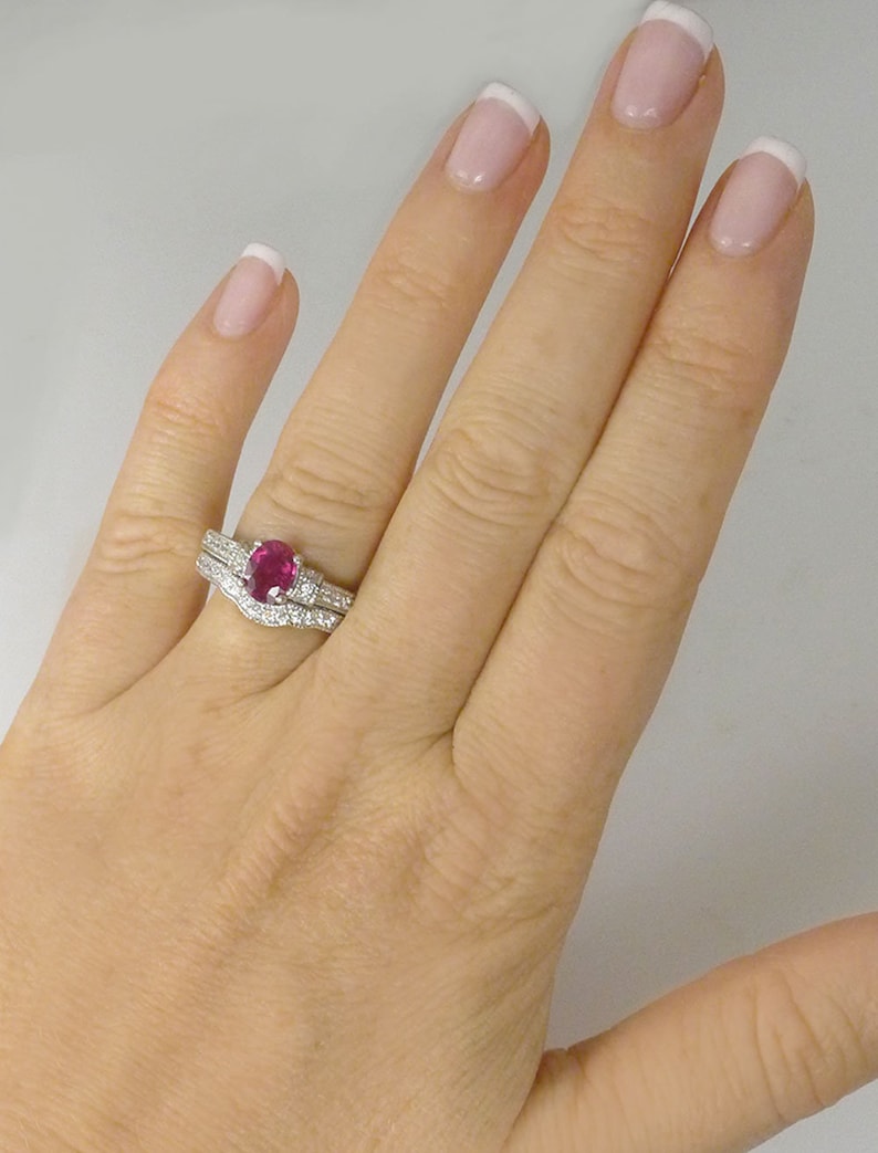 May include: A close-up of a woman's hand wearing a ruby and diamond engagement ring set. The ring features a large oval-shaped ruby surrounded by a halo of smaller diamonds. The band is also set with diamonds.