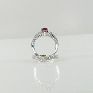 May include: A silver engagement ring set with a red ruby center stone and smaller diamonds on the band. The ring is paired with a matching wedding band with a diamond-encrusted design.