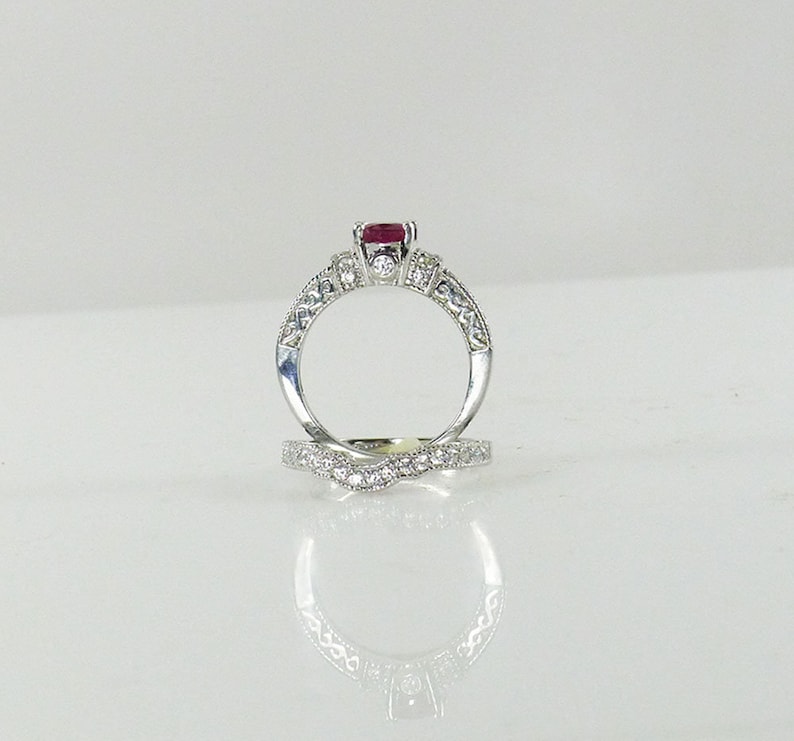 May include: A silver engagement ring set with a red ruby center stone and smaller diamonds on the band. The ring is paired with a matching wedding band with a diamond-encrusted design.