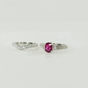 May include: A set of two silver rings with a red gemstone and clear stones. The engagement ring has a larger red oval gemstone and the wedding band has a wavy design.