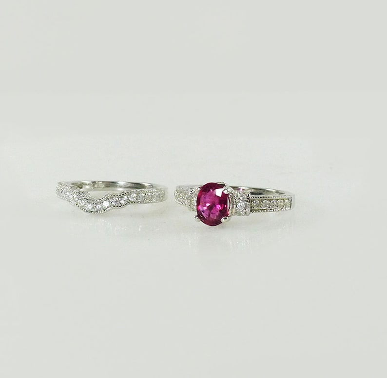 May include: A set of two silver rings with a red gemstone and clear stones. The engagement ring has a larger red oval gemstone and the wedding band has a wavy design.