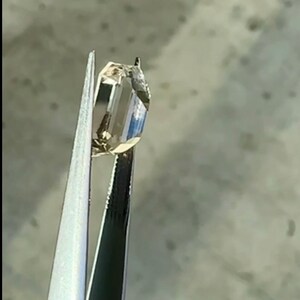 May include: A clear, faceted gemstone held in tweezers. The gemstone is a light brown color and has a rectangular shape.