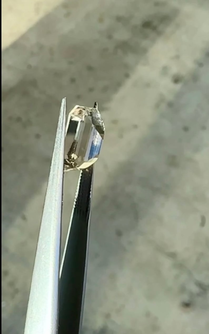 May include: A clear, faceted gemstone held in tweezers. The gemstone is a light brown color and has a rectangular shape.